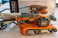 Photo of Ridgid heavy duty variable speed belt sander with dust collection bag, placed in front of its box showing features and branding.