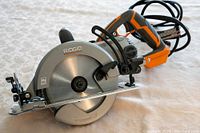 Power Ridgid 7 1/4 inch circular saw with orange and gray coloring, resting on a patterned surface with visible blade, handle, and cord.