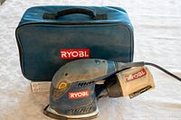 Ryobi corded corner sander with dust collection bag and blue carrying case on a white surface.