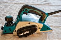 Side view of Makita KPO800 planer showing turquoise and off-white body, black adjustment knob, and handle with power cord.