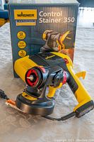 Photo shows Wagner Control Stainer 350 handheld paint sprayer next to the original box on a patterned surface.