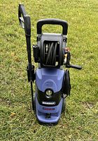 Simoniz blue and black pressure washer standing upright on grass showing front view with hose reel, spray wand and handle