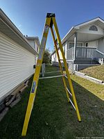 Photo showing a yellow 9 foot industrial Lite fiberglass step ladder standing open in grass beside a house.