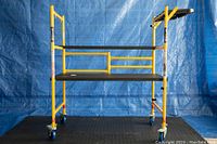 Full frontal photo of Metaltech scaffold showing the yellow metal frame, standing platform, side tray, and caster wheels on a black mat with blue backdrop.