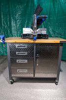 Photo shows a Mastercraft 12" dual bevel compound miter saw mounted on a metal storage center with four drawers and a diamond plate cabinet door. The storage center has a wood top and caster wheels for mobility.