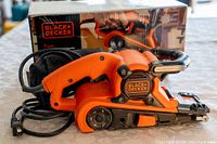 Orange Black & Decker belt sander next to its original box on a white patterned surface. Visible power cord, sanding belt, and product branding.