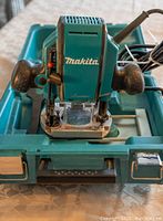 Makita electric router with two black handles and green carrying case beneath it, placed on a table.