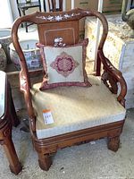 Full view of carved wooden Asian armchair with beige seat cushion and decorative pillow, showing inlaid floral designs on top rail and back panel.