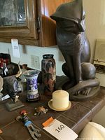 Large bronze cat statue with Egyptian-style scarf, small pottery vase with floral motifs, cloisonné vase, bronze bull bust, candles, and small decor items arranged on a tiled surface near wooden cabinetry.