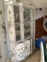 Angled front view of two-piece white lacquered chinoiserie china cabinet showing painted bird and floral motifs and glass display doors