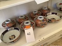 Seven covered bowls and two open serving bowls with detailed hand-painted Imari patterns, showcasing the full lot in the shelving unit.