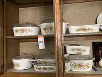 Full view of two shelves containing CorningWare ceramic cookware with French herb and vegetable pattern and clear glass lids.