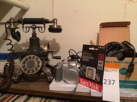 Front view of an ornate rotary dial landline phone and Canon PowerShot S2 camera with SanDisk memory card in package, also ACME Lite Super 8 camera light box and cables visible