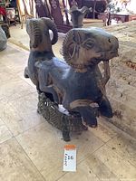 Front three-quarter view of two-headed ram sculpture on ornate metal stand placed on tile floor with background furnishings.