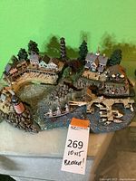 Full view of miniature village with small houses, lighthouse, docks, boats, and rocky cliffs beside water. Note village is broken into pieces.