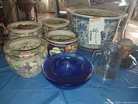 Five Asian ceramic planters with various traditional designs, a clear glass cylindrical container, a bust figurine of Nelson Mandela, and a blue glass ashtray on a table.