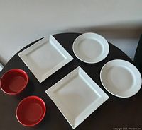 Photo showing all six items laid out on a round black table: 2 red bowls, 2 white square plates, and 2 white round plates.