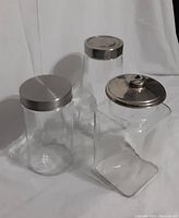 Three clear glass canisters with metal lids displayed on a white background showing different shapes and lid types.