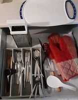 Photo showing cutlery tray with forks, knives, spoons and teaspoons, wooden-handled utensils, two red oven mitts, metal cheese grater, white cutting board and white plastic spoon with wooden handle.