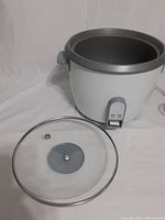 Electric rice cooker with glass lid placed beside it showing the inside of the cooker and pot removal.