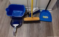 Blue cleaning bucket, non-standing dustpan with white brush, yellow broom, blue standing dustbin with handle and black brush, broom with black handle all placed on floor