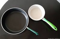Two frying pans viewed from above on a black surface, showing the Revere Ware pan with black non-stick interior and the smaller Dupuis Aubecq pan with white ceramic lining.
