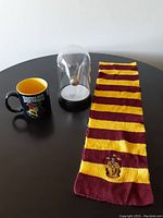 Photo of all three items on a black round table: Gryffindor scarf, Golden Snitch glass cloche light, Hogwarts crest mug