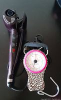 Conair Infinity Pro Curling Iron placed next to portable luggage scale with leopard print design on black background