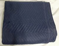 Folded dark blue quilted moving blanket showing multiple layers and stitching detail. Visible small tear near edge.