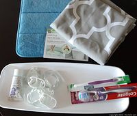 New light blue plush bathtub mat, new fabric gray and white geometric shower curtain, white tray with small bottle of bath oil, clear shower curtain rings, and two toothbrushes in original packaging on table.