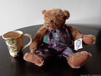 Photo showing the Ganz oversized thick mug with hand-edged ridges and grape and vine decoration next to the Chuckles teddy bear in plaid overalls, seated on a round table.