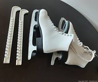 Pair of white leather ice skates with black soles and silver blades next to two white plastic skate guards on a black surface