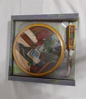 Front view of the boxed wine art round cheeseboard and cutter set showing the colorful engraved wine table setting design through clear packaging.