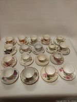 Full view of all 18 vintage floral ceramic tea cups arranged in three rows with their matching saucers, showing various delicate floral patterns and gold trim detailing.