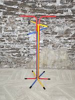 Front view of the red, yellow, and blue tubular metal clothing/coat rack against a rough stone wall and tile floor.