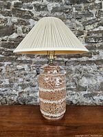 Full view of the ceramic lamp with pleated beige shade against a stone wall background.