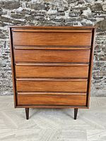 Front elevation of the dresser showing five drawer fronts and tapered legs against stone wall.