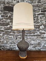 Full view of the lamp showing the textured brown ceramic base and tall beige fabric shade against a stone wall background.