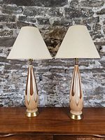 Full view of two lamps side by side on wood surface against brick wall
