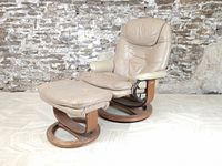 Front angle showing beige leather lounge chair with plush cushioning and matching footstool on circular wooden bases, leather slightly worn.