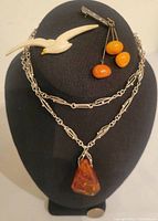 Three jewelry pieces displayed on black stand: amber pendant necklace, amber pin with three beads, and cream colored resin bird brooch.