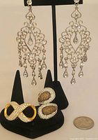 Front view of silver filigree rhinestone chandelier earrings with teardrop dangles and two rhinestone rings on black stands with quarter for scale