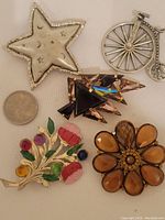 Photo showing all five brooches arranged near a quarter for size reference: silver star, vintage bicycle, amber flower, colorful flower, and geometric kite-shaped brooches.