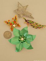 Overview of the four brooch pins including rhinestone star, butterfly with colorful rhinestones, large green flower, and peacock, shown with a quarter for size reference