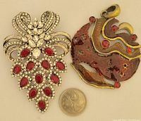 Two jewelry pieces: a large silver-tone rhinestone brooch and a brass enamel pendant with red rhinestones, shown next to a coin for scale.