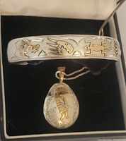 Top-down photo showing the silver-tone adjustable bangle with raised 12k gold filled figures including birds, a lizard, a footprint, the pendant with similar figures and a sun motif, resting in a black jewelry box.