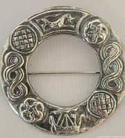 Front view of the circular silver pin showing embossed Scottish-themed motifs including a lion, thistles, and Celtic knot patterns.