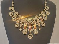 Front view of the large bib necklace featuring gold tone metal and multiple clear and iridescent glass stones with amber rectangular accents