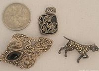 Three pieces of silver and marcasite jewelry including a floral brooch with black enamel and marcasite stones, a marcasite pendant with a black stone, and a dog brooch with marcasite accents shown next to a coin for scale.