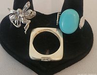 Three silver rings displayed together on black ring holder. Includes fairy figure ring on the left, square band ring in center, and turquoise stone ring on right.